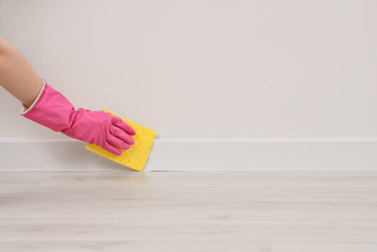 Person in pink rubber glove cleaning white baseboard with yellow sponge, home maintenance and hygiene