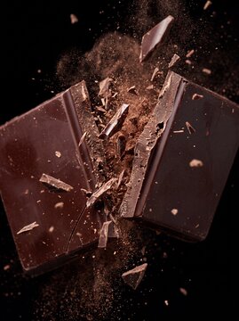 Exploding dark chocolate bar with flying shards and cocoa powder on black background