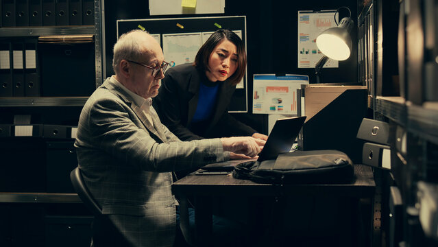 Law agents studying old classified information from police archives folders, looking for clues and crime trends. Partners accessing additional archived resources in incident room. Camera B.