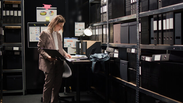 Caucasian policewoman conducts investigative legal work with confidential papers, working on a case. Examining witness statements on laptop, detective cross-references with files from archive shelves.