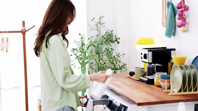Woman kitchen washing machine detergent home appliance casual lifestyle cleaning domestic modern wooden countertop plant coffee maker cup dish rack plate bowl towel garlic bright natural light