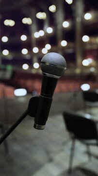 A microphone is positioned onstage in a theater. The background shows rows of empty seats and lights shining above. People gather outside in anticipation of the performance.