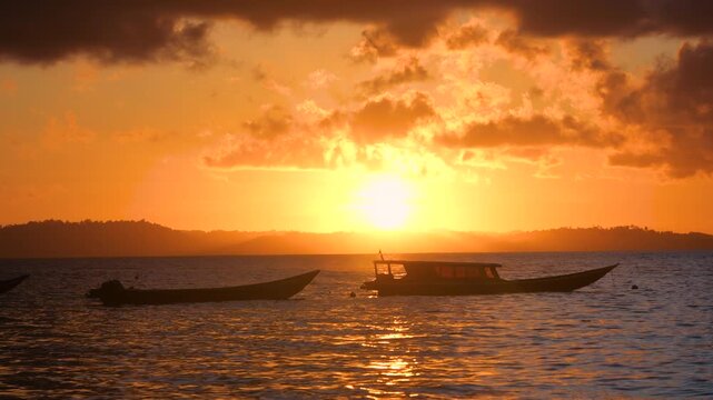 SILHOUETTE, LENS FLARE: Two traditional boats rest beneath a fiery tropical sky as sun sinks over Indian Ocean. Golden reflections shimmer across calm sea during a peaceful evening at Mentawai Islands
