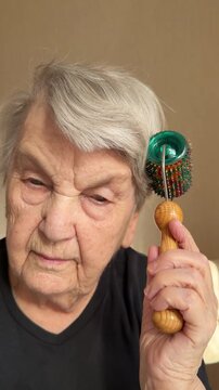 Elderly Caucasian grandmother scalp massage close-up, seated indoors using green magnetic needle roller on grey hair, gentle