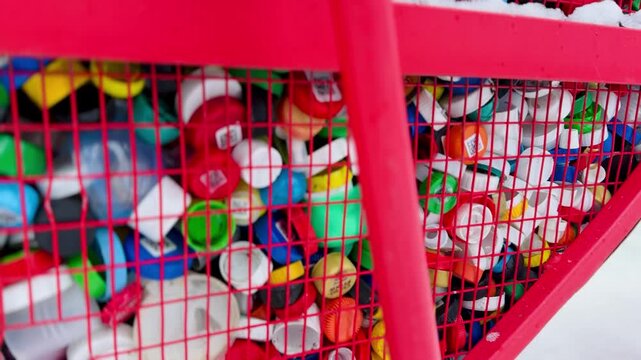 Close-up view of a red wire mesh basket containing a pile of assorted colorful plastic bottle caps. Shot from a slightly low angle with soft side lighting
