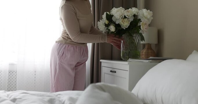 Woman putting vase with beautiful peonies on bedside table and turning on lamp at home, closeup