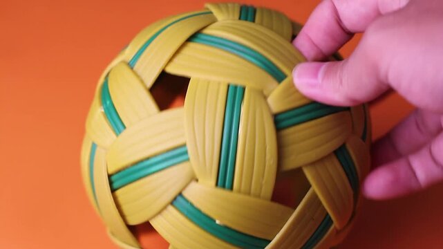 Close-up shot of a person's hand holding and showcasing a traditional woven sepak takraw ball against a solid bright orange background.