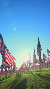 Vertical Screen: USA Flag September 11 memorial tribute displays hundreds of American flags waving on green grass field during golden hour with sun flares and long shadows across the lawn.