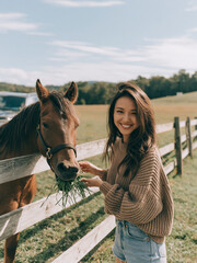Obraz premium A smiling young woman in a knitted sweater stands by a wooden fence and feeds fresh green grass to a brown horse in a sunny field