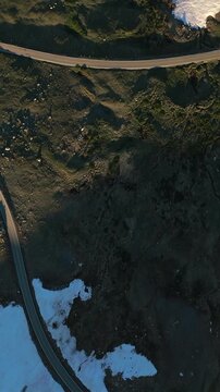 Vertical Screen: Beartooth Highway in Montana USA showcases curved asphalt road cutting through rugged alpine terrain with patches of melting snow and sparse vegetation during golden hour light.