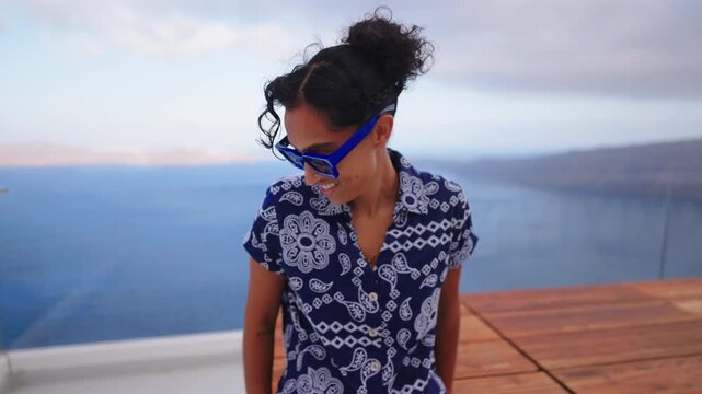Smiling Indian woman wearing blue sunglasses and patterned shirt sits on wooden terrace overlooking calm sea in Santorini Greece with mountains and cloudy sky in background during evening.