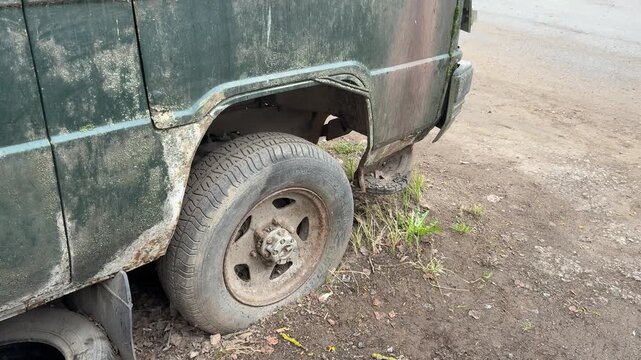 Abandoned dark green minibus with punctured tire, close-up of muddy wheel and spare rim tucked under chassis, heavy rust patches