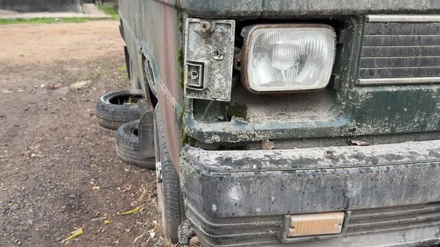 Rusted dark green minibus broken headlight, parked in gravel lot during spring, flaking paint and moss covering corroded body