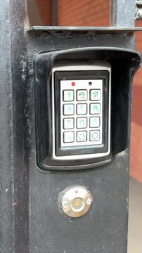 Analog intercom keypad on iron gate, weathered black frame, green backlit numeric buttons, round scanning key below keypad, brick