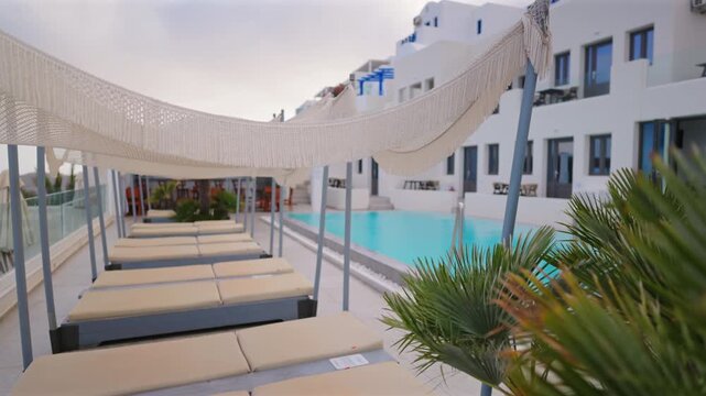 Elegant resort poolside in Santorini Greece featuring rows of beige sun loungers under woven canopy shades beside turquoise swimming pool with traditional white Cycladic buildings and palm fronds.