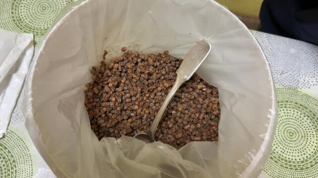 White bucket filled bee pollen granules on patterned tablecloth, metal scoop resting inside bucket, market vendor display