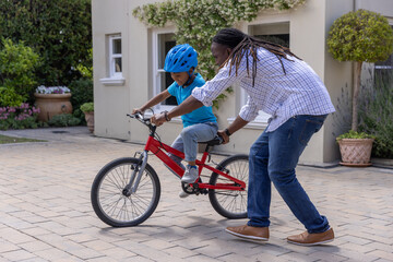 Fototapeta premium African American man steadying boy wearing blue helmet on red bicycle on driveway, in plaid shirt