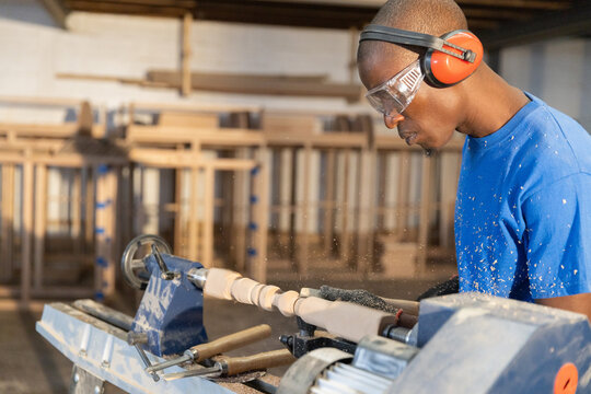 Wood lathe shaping cylindrical spindle in carpentry workshop, with scattered shavings on shelves