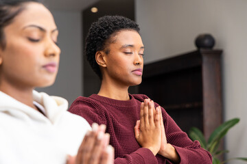 Fototapeta premium Indian, African American women meditating with hands in prayer at home, maroon sweater white hoodie