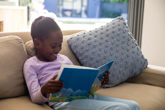 African American girl child sitting on sofa at home, holding blue picture book, wearing purple top