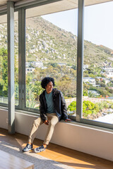 Naklejka premium African American man in 20s sitting on wide windowsill by glass walls, sunlight on jacket