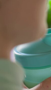 Vertical video: Gripping toddler raising teal cup with spout in kitchen for thirst, in striped top