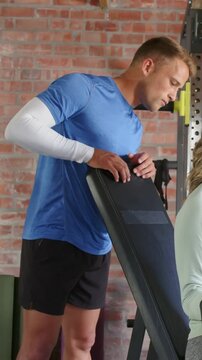 Vertical video: Adjusting trainer securing adjustable bench at gym after partner cue, in blue shirt