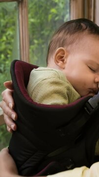 Vertical video: Entering parent cupping and stroking sleeping baby in carrier by window, soothing