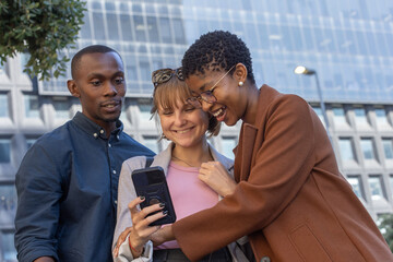 Diverse friends leaning in urban plaza, sharing smartphone with dark case, wearing blazer and coat © wavebreak3