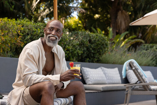 Mature man wearing beige shirt and shorts, sitting poolside holding tall glass with strawberry