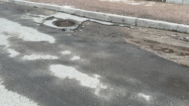 Worn asphalt road with potholes showing curbside emergency lane, gravel shoulder, standing puddles, cracked surface, localized