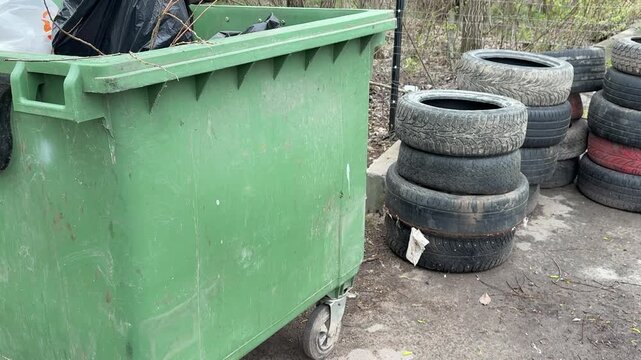 Green dumpster and stacked tires on gritty roadside during spring day, worn rubber wheels piled beside municipal container