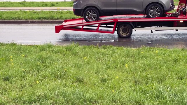 Close-up passenger car on red flatbed tow truck, spring roadside scene with wet pavement and puddles, green grassy verge dotted