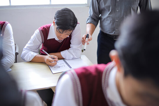High School Student Writing in Notebook During Class Exam While Teacher Guides Him