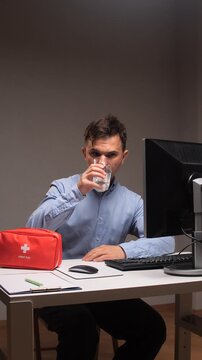 Vertical video man sitting at office desk, taking a pill with a glass of water, struggling with headache and stress from long hours and deadlines while working on computer paperwork