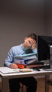 Vertical video overworked male employee experiencing exhaustion and fatigue while leaning on a towering stack of files and documents next to a computer at a desk
