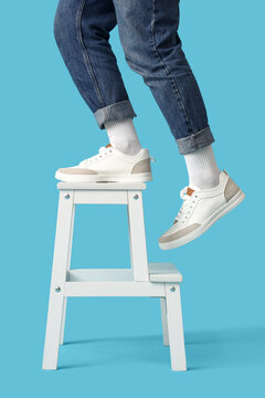 Person in jeans and white sneakers stepping up on a white step stool, isolated on a bright blue background