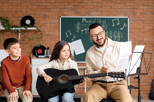 Music teacher guiding young girl playing acoustic guitar with boy observing in classroom setting, the teacher holds sheet music, demonstrating notes to the focused student