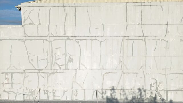A drone shot showing a large, weathered white concrete wall, with cracks and patches of plaster on the exterior of the old building. 
