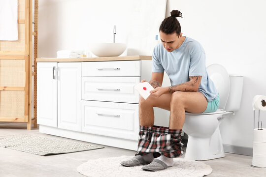 Young man experiencing hemorrhoids or digestive issues, holding toilet paper with blood stain in bathroom, he appears to be experiencing discomfort or pain