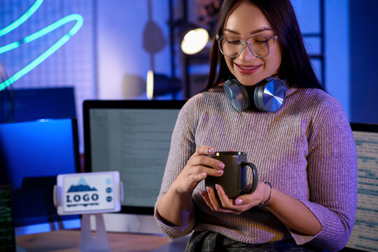 Beautiful young female programmer with cup of coffee near her workspace in modern office at night