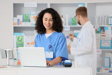 Smiling female pharmacist in blue scrubs using laptop at pharmacy counter with male colleague, a...