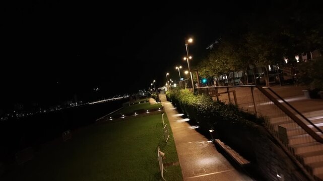 Empty paved walkway and green grass illuminated by streetlights along lake at night in Malgrate, Italy. Aerial, spin shot, copy space