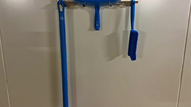 Professional cleaning equipment including a blue floor scraper, dustpan and brushes hanging on a wall rack inside a commercial space