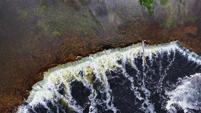 Overhead close drone shot of weir edge creates an abstract pattern of white foam on dark river. Kuldīga, Latvia.