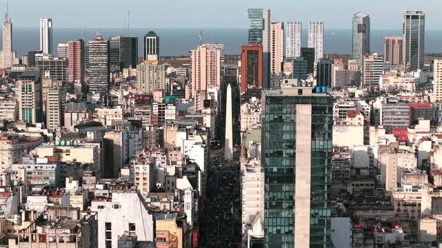 Avenida Corrientes leads directly to the Obelisk of Buenos Aires through the dense city centre skyline, with modern office towers and the R&iacute;o de la Plata visible in the distance, slow forward push-in