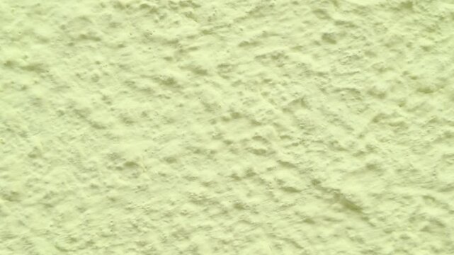 Outdoor panning shot captures the rough bumpy surface texture of an industrial plaster building wall painted in a pale light yellow cream tone on a bright overcast daytime afternoon.