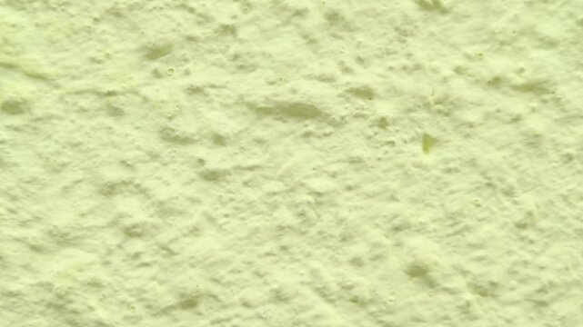 Outdoor panning close up shot captures the rough bumpy surface texture of an industrial plaster building wall painted in a pale light yellow cream tone on a bright overcast daytime afternoon.
