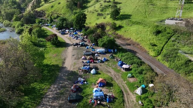 Homeless encampment with colorful tents and blue tarps spreads along dirt tracks beside river beneath lush green hillside on sunny spring day in Roseburg, Oregon, USA. Aerial drone view.