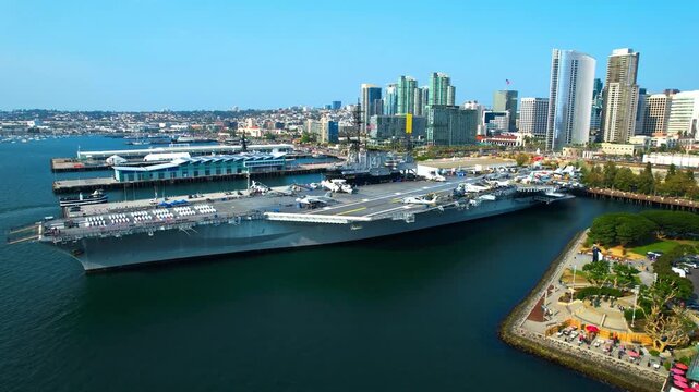 Cinematic high-angle drone shot looking down at the USS Midway flight deck and downtown skyscrapers.